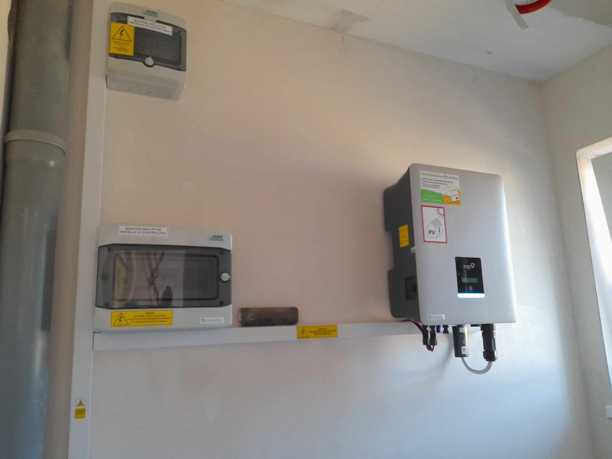 Szara skrzynka elektryczna i falownik PV FoxESS zamontowane na ścianie. Widoczne kable i ostrzeżenia. Instalacja w pomieszczeniu.