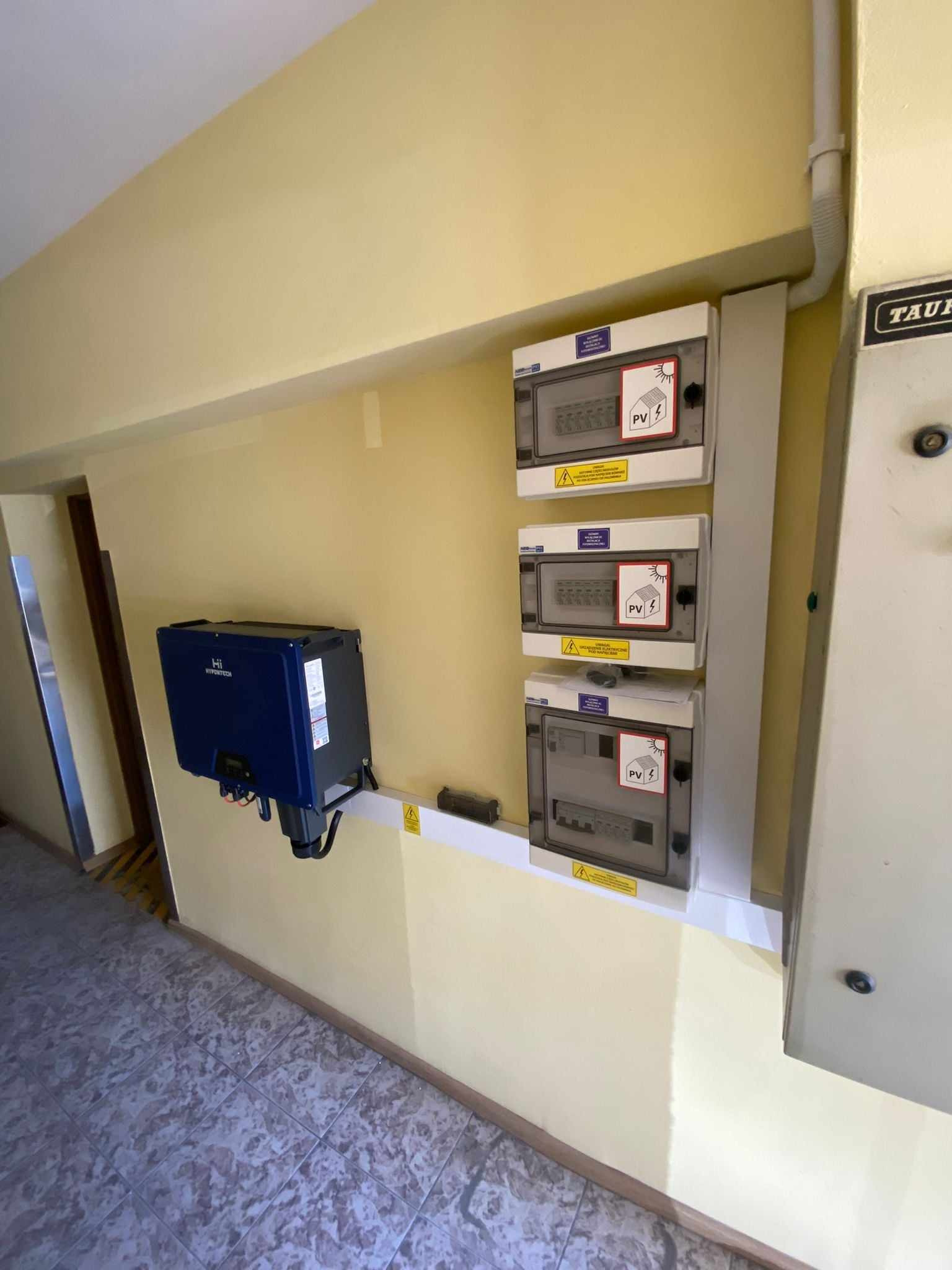 Niebieski inwerter fotowoltaiczny Hi Hypertech na żółtej ścianie obok trzech skrzynek bezpiecznikowych z naklejkami PV. Instalacja wewnątrz budynku.