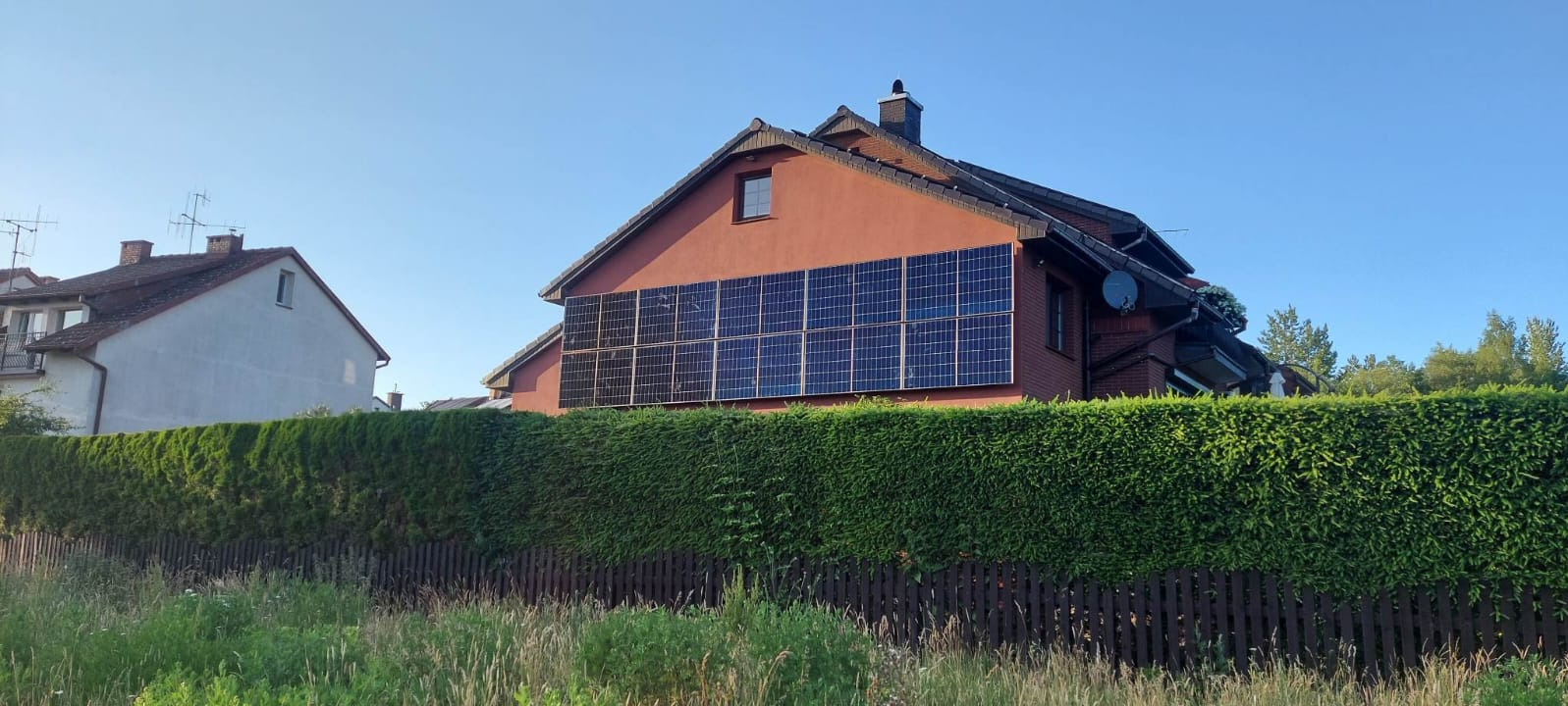 Czerwony dom jednorodzinny z panelami fotowoltaicznymi na ścianie szczytowej, widoczny zza żywopłotu i drewnianego płotu. Błękitne niebo.