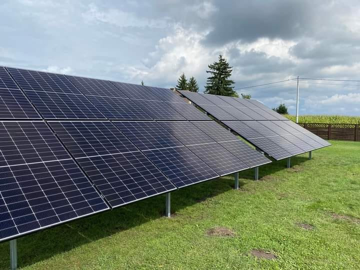 Dwa rzędy paneli słonecznych na trawniku, zamontowane na metalowych podporach. W tle pole kukurydzy i pochmurne niebo.