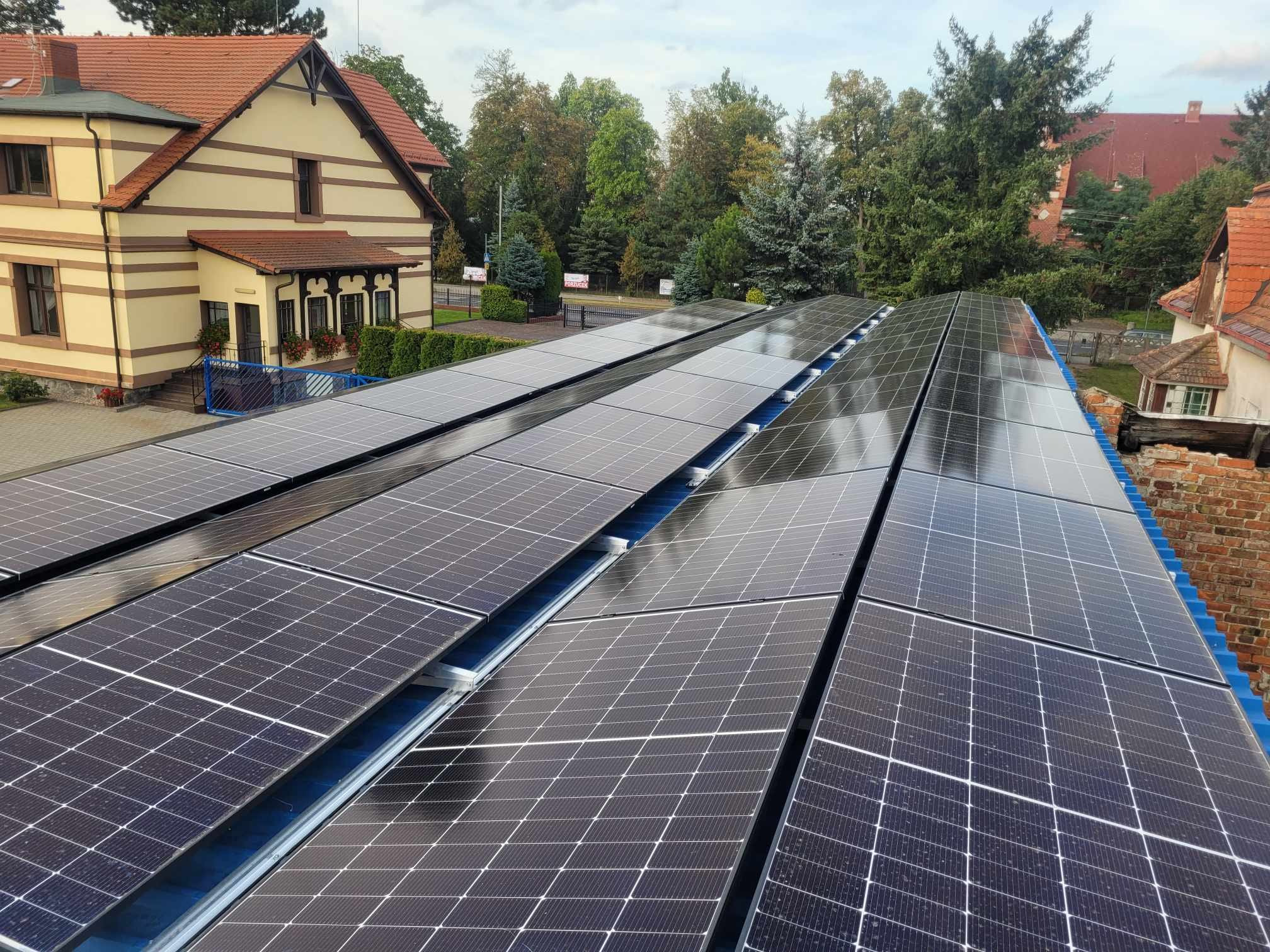 Rząd paneli słonecznych na dachu budynku z czerwonej cegły, w tle drzewa i budynki. Widok z góry, lekki kąt. Błękitne niebo z delikatnymi chmurami odbija się w panelach.