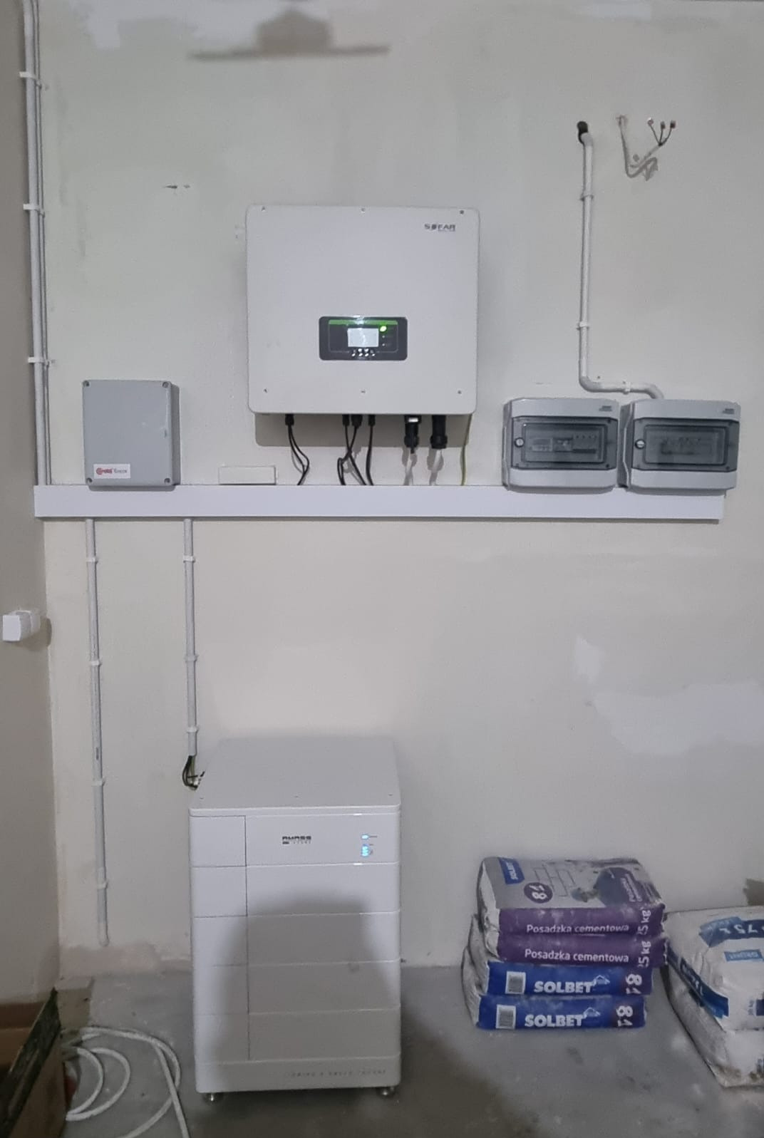 Biały inwerter i magazyn energii Dyness zamontowane na ścianie, widoczne kable, obok worki z posadzką cementową Solbet. Instalacja w surowym pomieszczeniu.