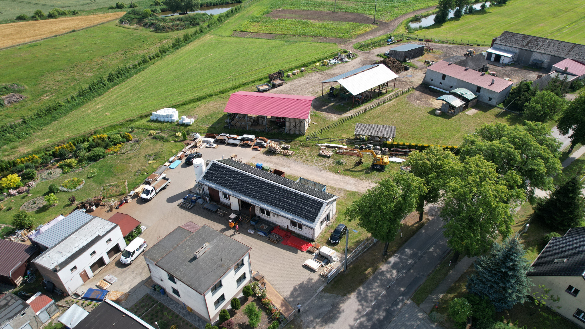 Widok z lotu ptaka na budynek z panelami słonecznymi na dachu, otoczony zabudowaniami gospodarczymi i polami uprawnymi w słoneczny dzień.