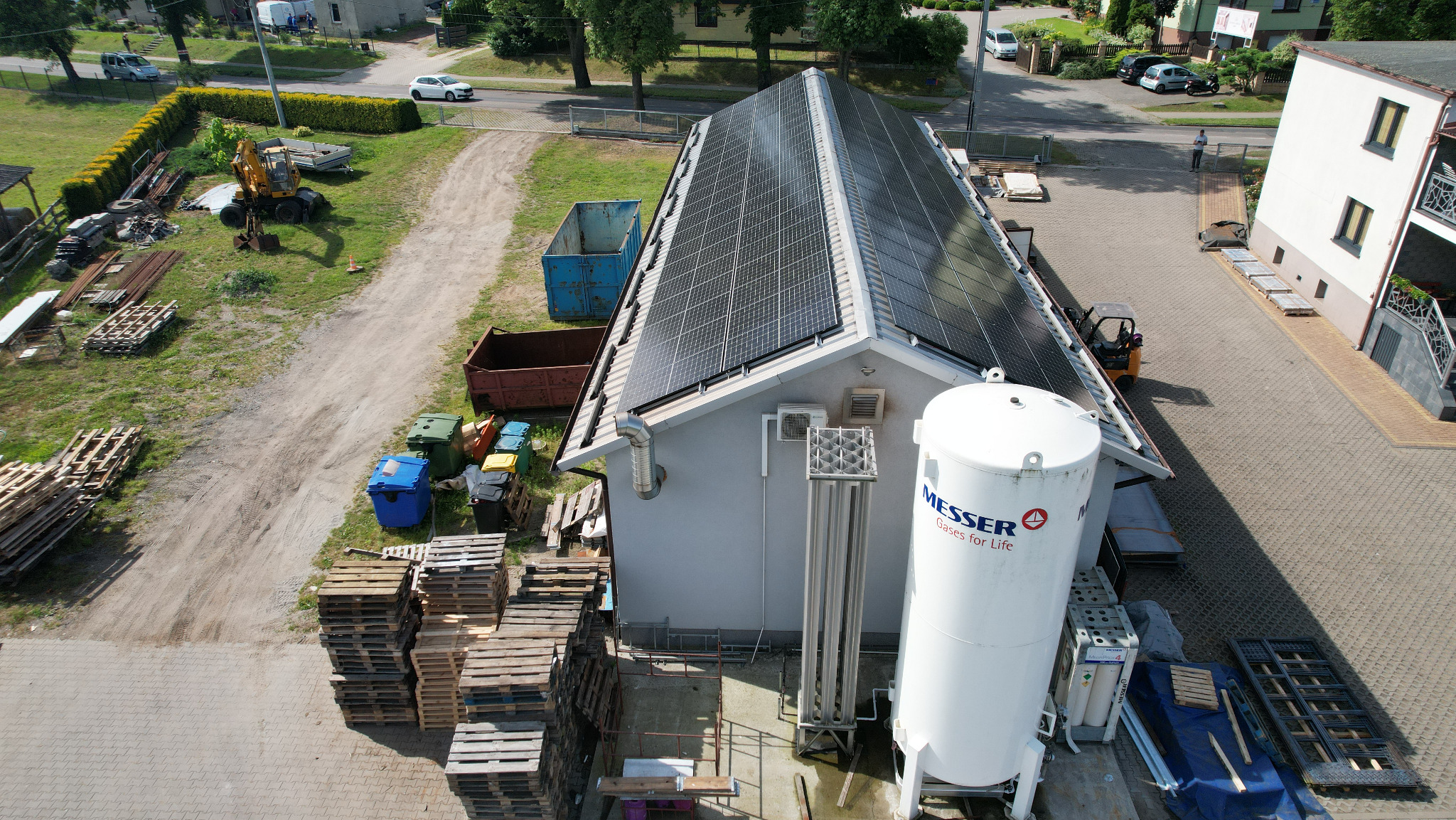 Instalacja paneli na dachu budynku przemysłowego, widok z góry. Obok zbiornik Messer i palety. W tle plac z koparką i kontenerami.