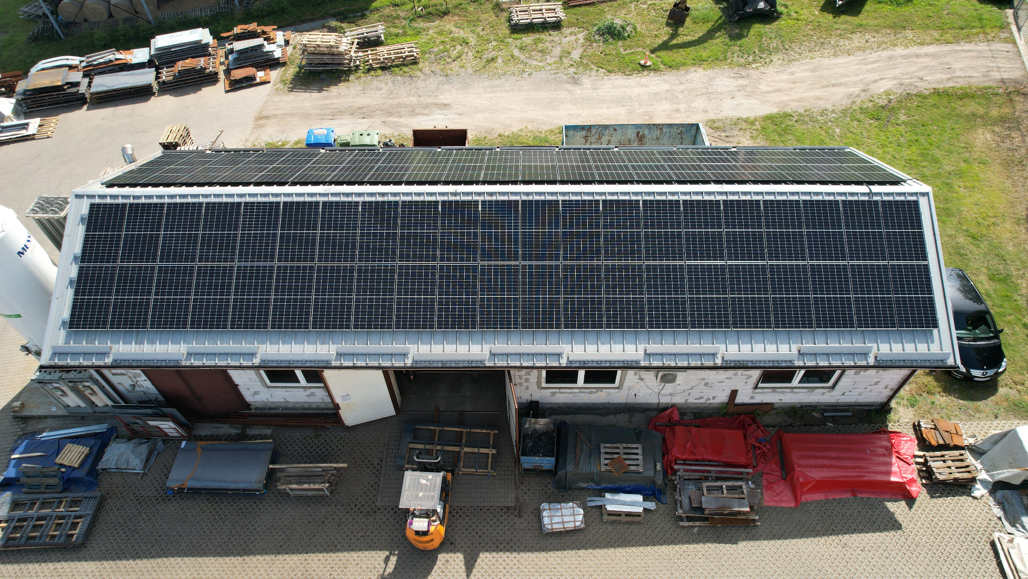 Widok z góry na budynek z panelami słonecznymi na dachu, obok zaparkowany samochód, w tle palety i inne materiały budowlane. Na dole wózek widłowy.