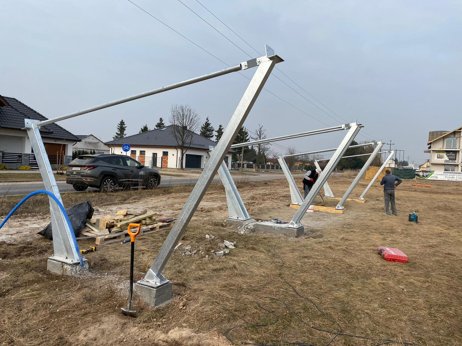 Montaż konstrukcji wsporczej pod panele słoneczne na fundamencie betonowym. Widoczne narzędzia i pracownicy. W tle domy jednorodzinne i pochmurne niebo.