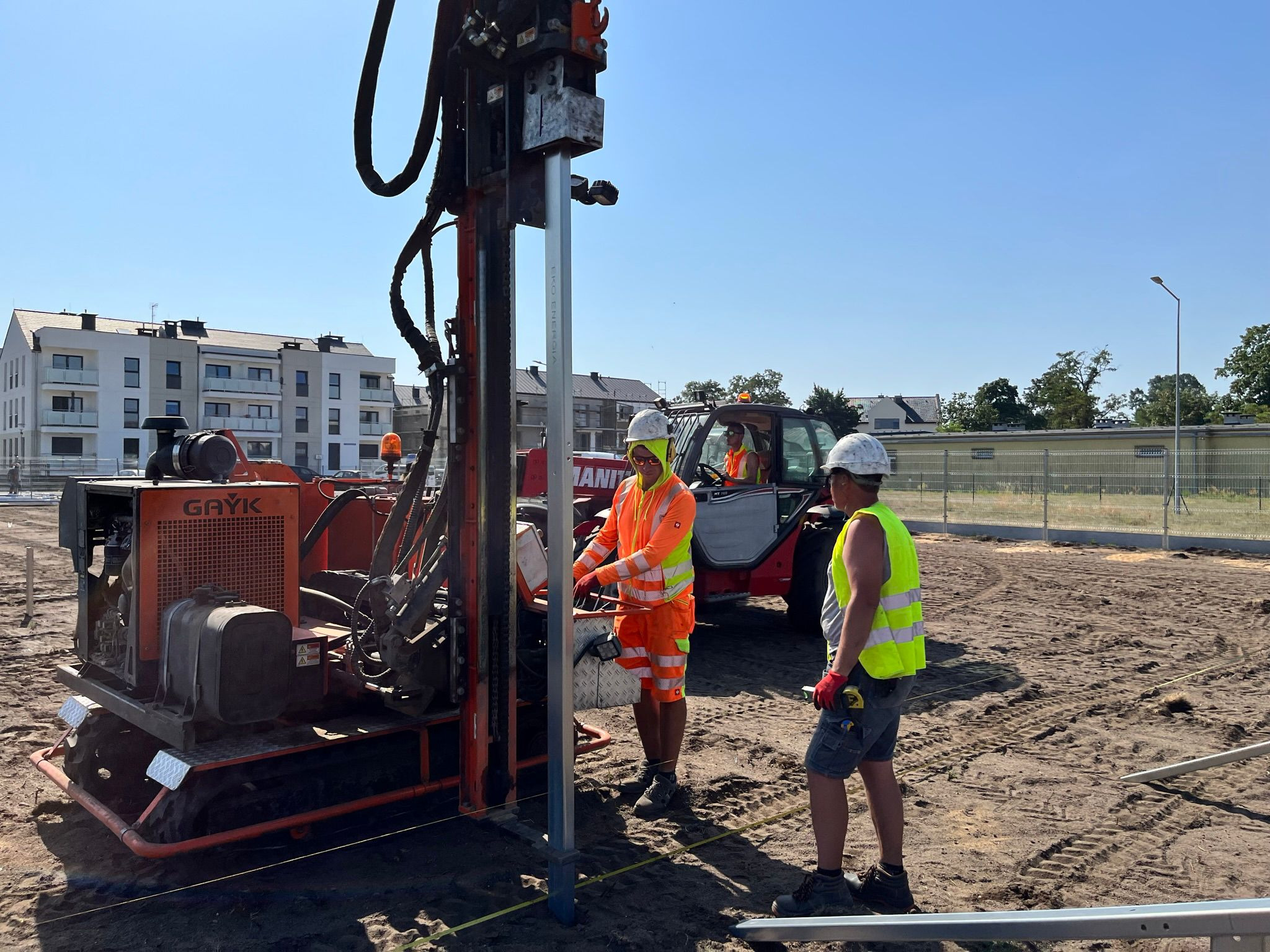 Pracownicy w odblaskowych ubraniach obsługują maszynę wiertniczą Gayk na placu budowy, w tle budynek mieszkalny i maszyna budowlana Manitou.