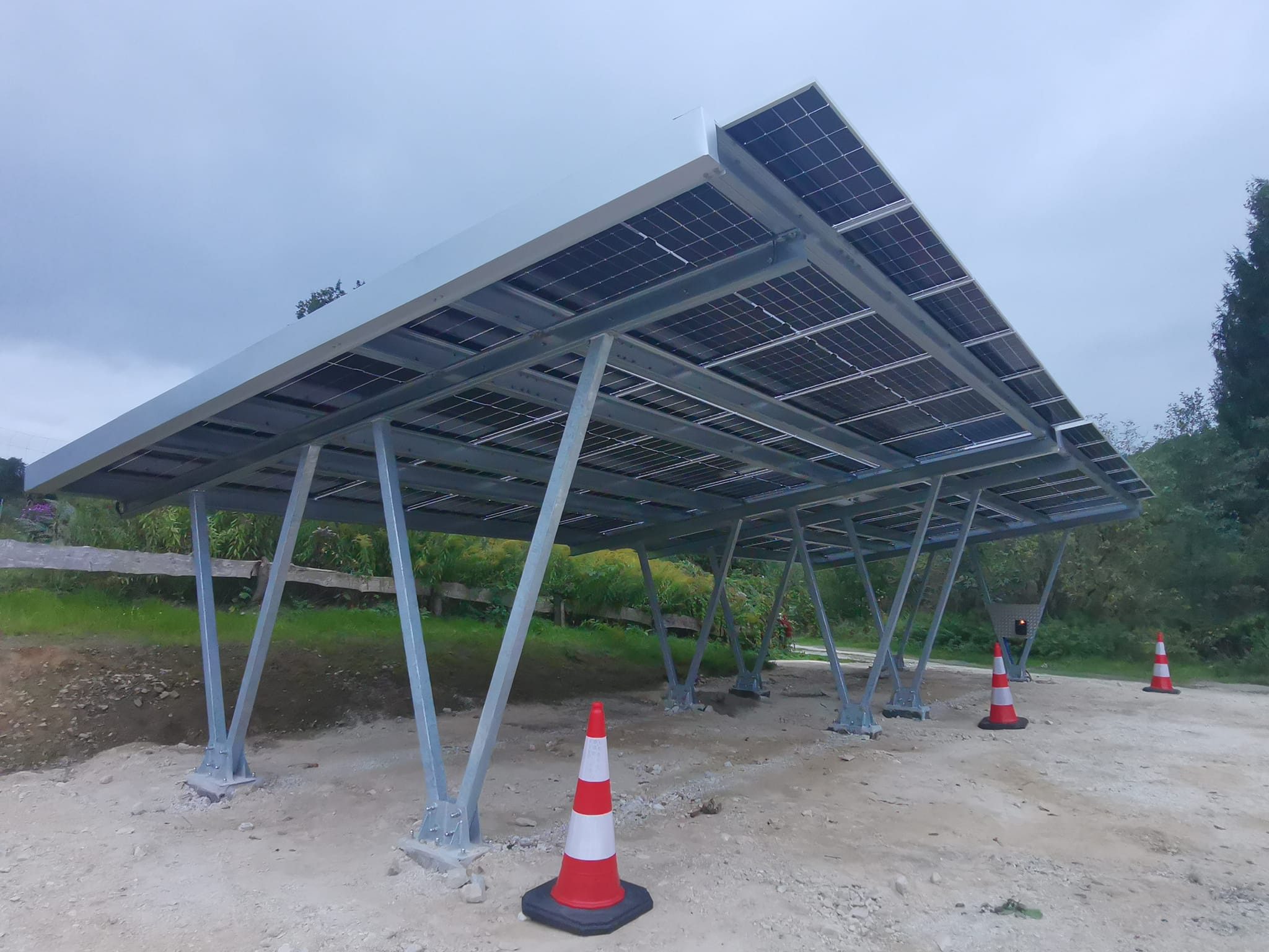 Zadaszenie parkingowe z panelami słonecznymi, konstrukcja stalowa, widoczne słupki i panele, teren budowy z pachołkami. Pochmurne niebo w tle.