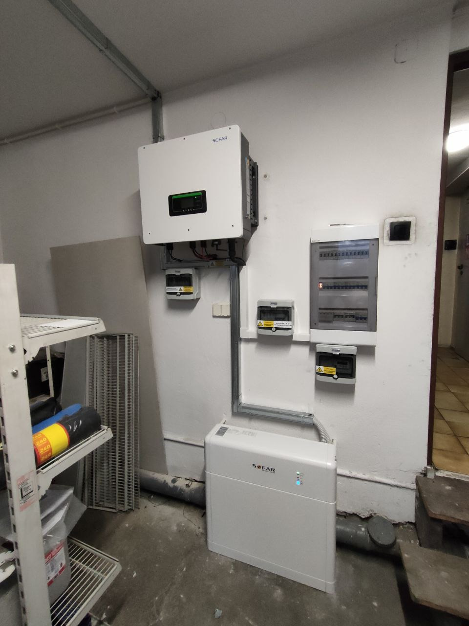 Biały inwerter Sofar i akumulator energii w pomieszczeniu, z widoczną rozdzielnicą elektryczną i przewodami. Instalacja fotowoltaiczna w garażu.