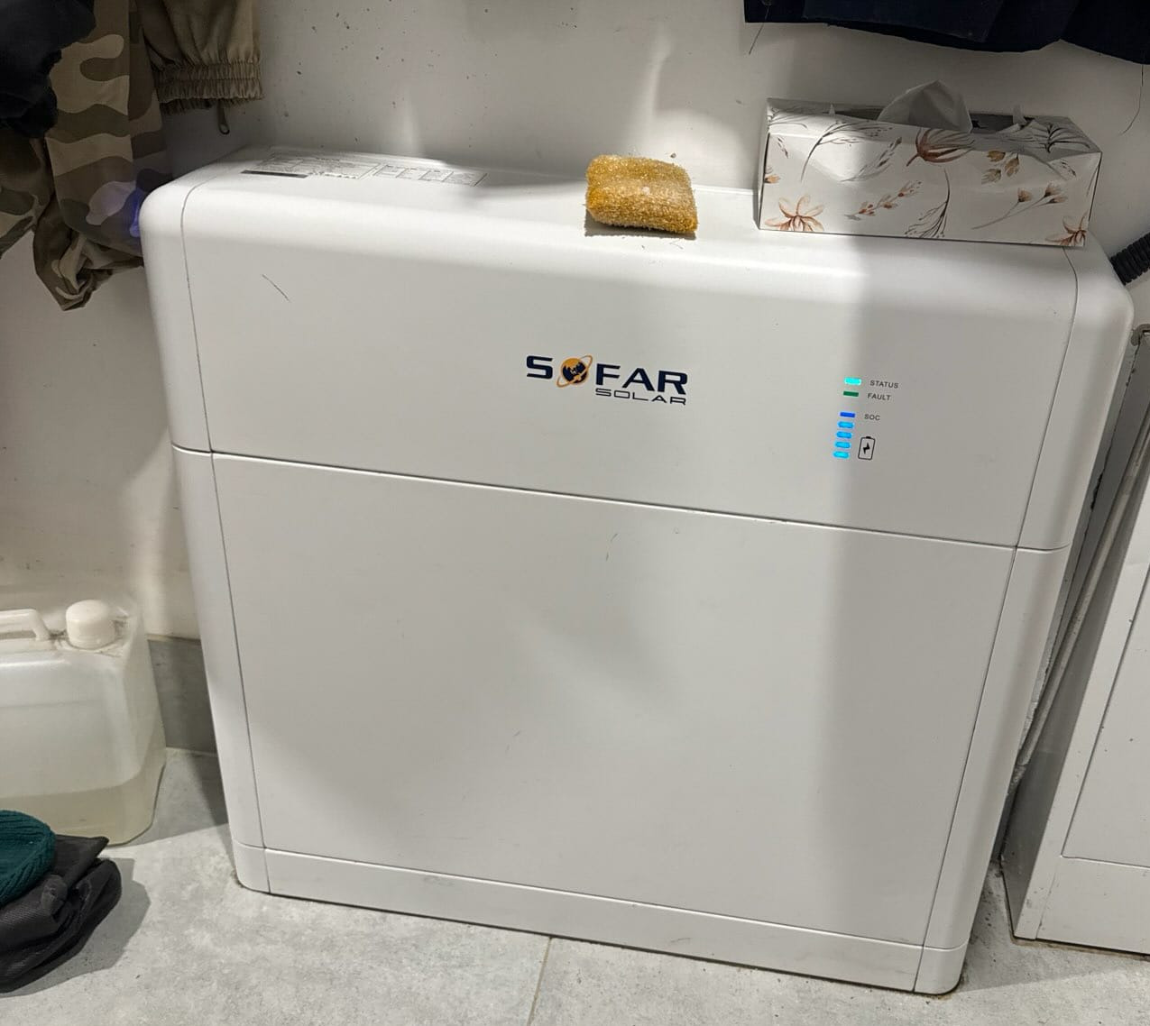 Biały magazyn energii SOFAR Solar z diodami LED stanu, umieszczony w pomieszczeniu. Na górze pudełko chusteczek i gąbka. Widoczne ubrania i pojemnik z płynem.