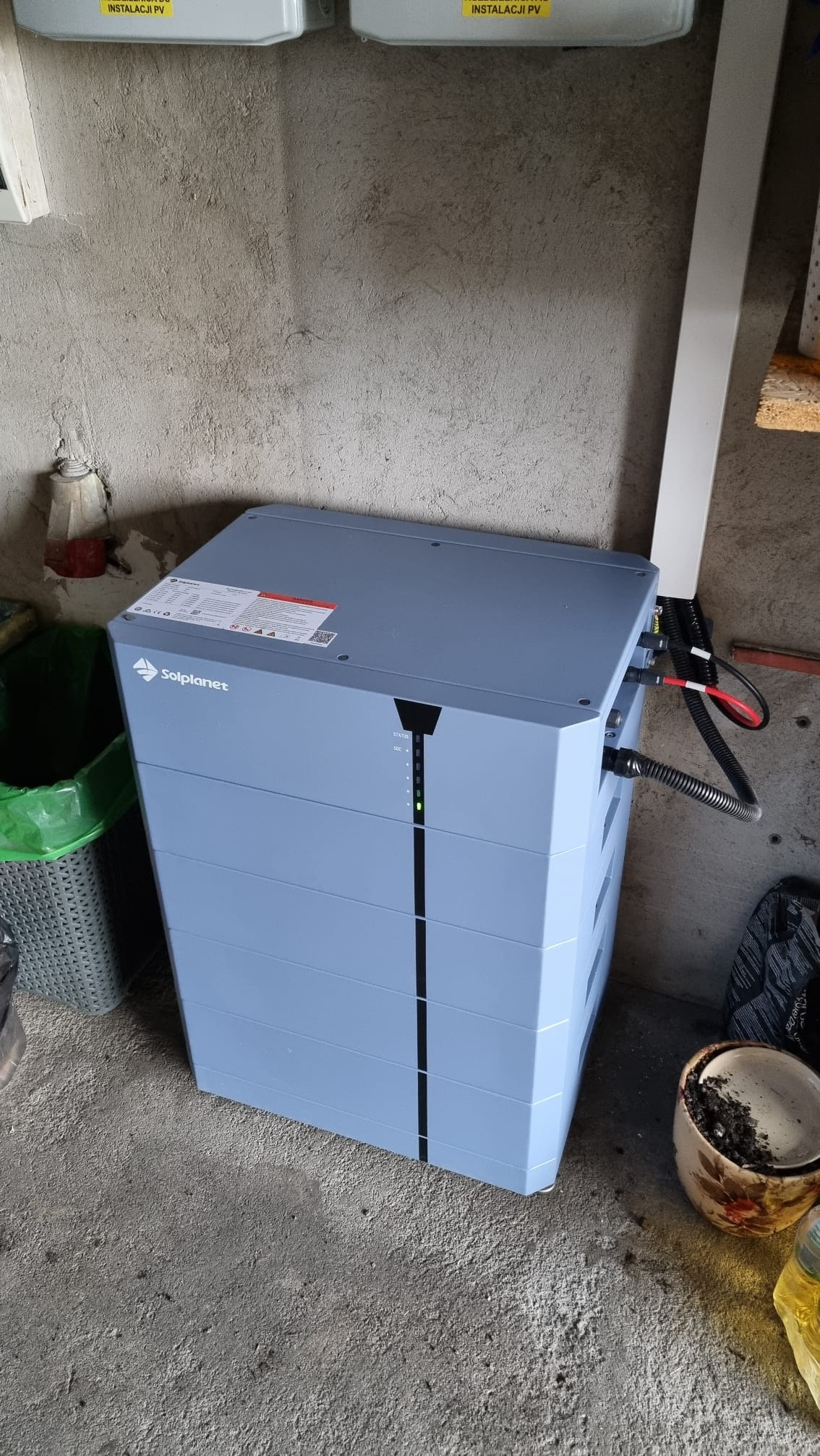Szary magazyn energii Solplanet w surowym wnętrzu, widoczne kable i wskaźnik naładowania. Obok kosz i doniczka, ściana z widoczną instalacją PV.