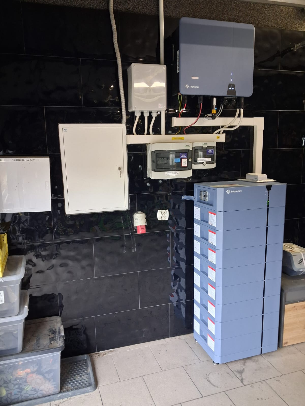 Instalacja magazynu energii Solplanet na tle czarnej, błyszczącej ściany z płytek. Widoczne skrzynki elektryczne, okablowanie i gniazda.