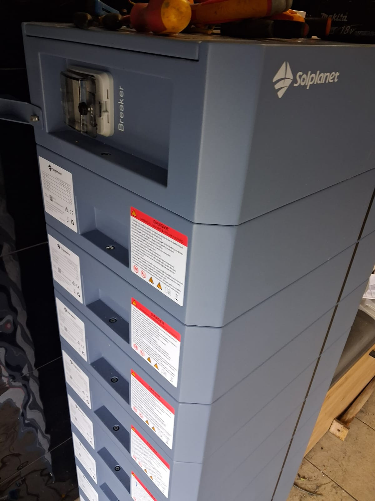 Szary, modułowy magazyn energii Solplanet z widocznym wyłącznikiem i naklejkami ostrzegawczymi. Na górze narzędzia, w tle błyszcząca powierzchnia i fragment palety.