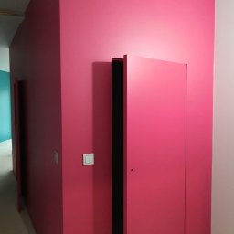 Izol Szczecin Andrzej Jobczyk - Różowa ściana z ukrytymi drzwiami i włącznikiem światła. Minimalistyczny design wnętrza, świeżo po malowaniu. Widoczny fragment podłogi zabezpieczony folią.