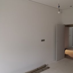 Izol Szczecin Andrzej Jobczyk - Świeżo pomalowane, białe wnętrze z widocznymi rolkami malarskimi przy ścianie i otwartymi drzwiami do kolejnego pomieszczenia. Minimalistyczny wystrój, surowy stan wykończenia.