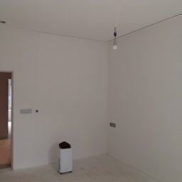 Izol Szczecin Andrzej Jobczyk - Wnętrze pokoju po malowaniu ścian na biało, widoczny oczyszczacz powietrza, surowa żarówka i narzędzia malarskie na podłodze. Świeżo pomalowane ściany.