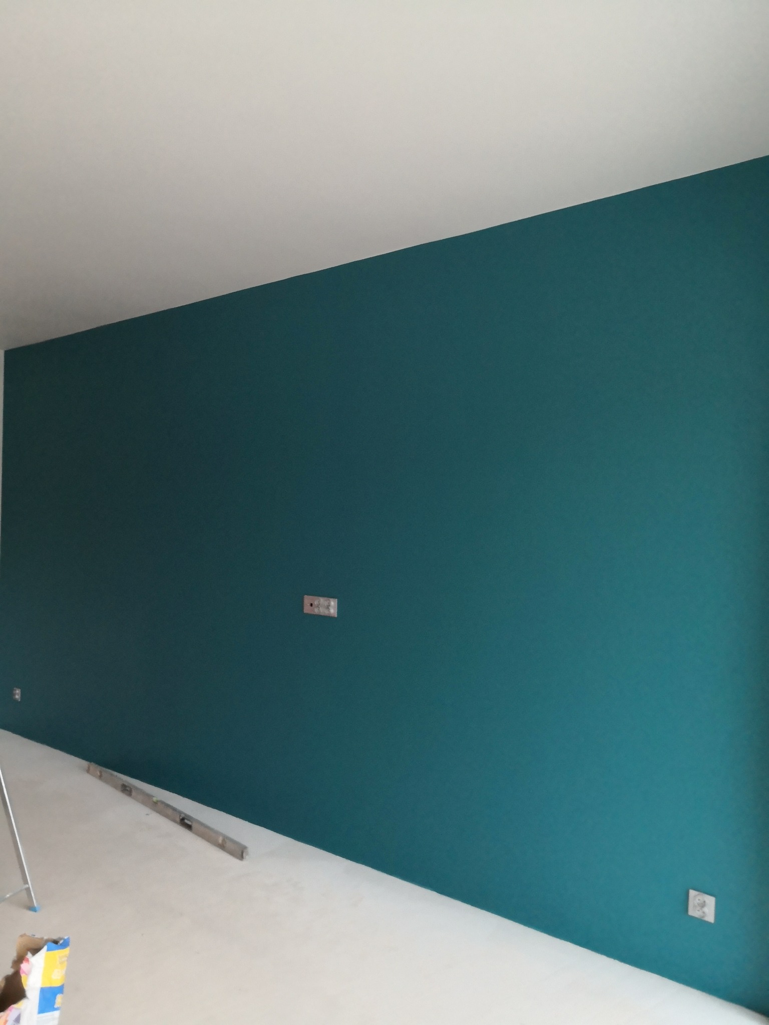 Ściana pomalowana na turkusowo, widoczne gniazdka elektryczne, poziomica na podłodze. Świeżo pomalowane wnętrze w trakcie wykańczania.