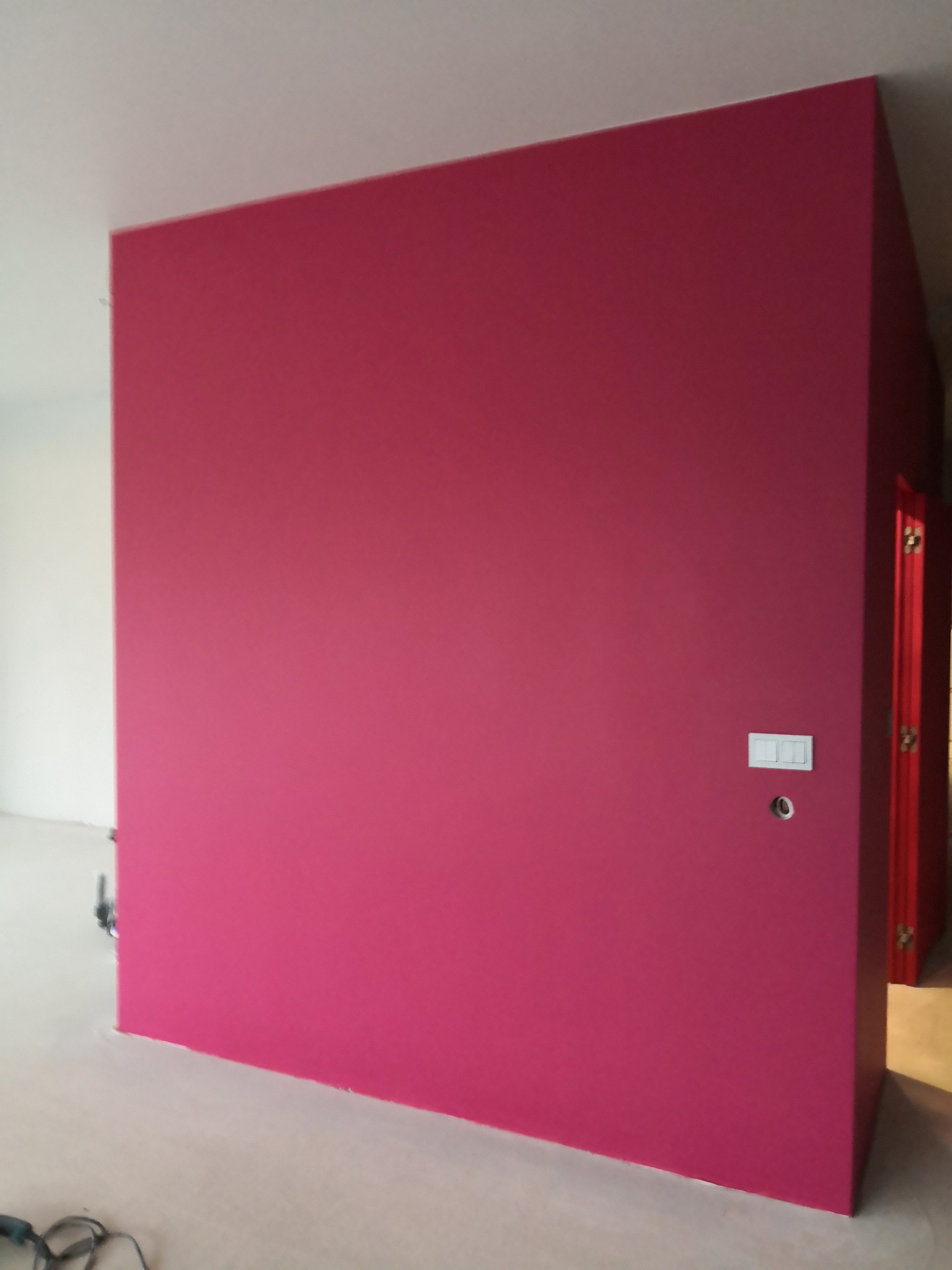 Ściana pomalowana na intensywny róż, z widocznym włącznikiem światła i otworem na gniazdko. W tle fragment białej ściany i czerwone drzwi.