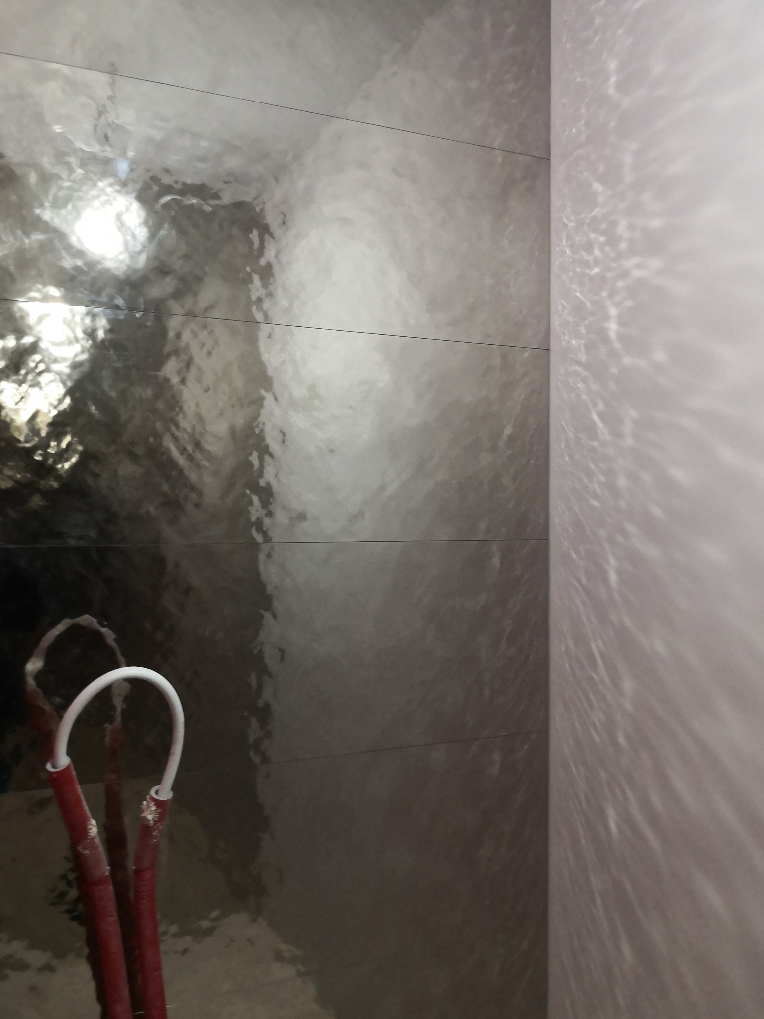 Położone, błyszczące, szare płytki na ścianie w łazience. Widoczne rury instalacji wodnej. Efekt lustrzanego odbicia światła na powierzchni płytek.