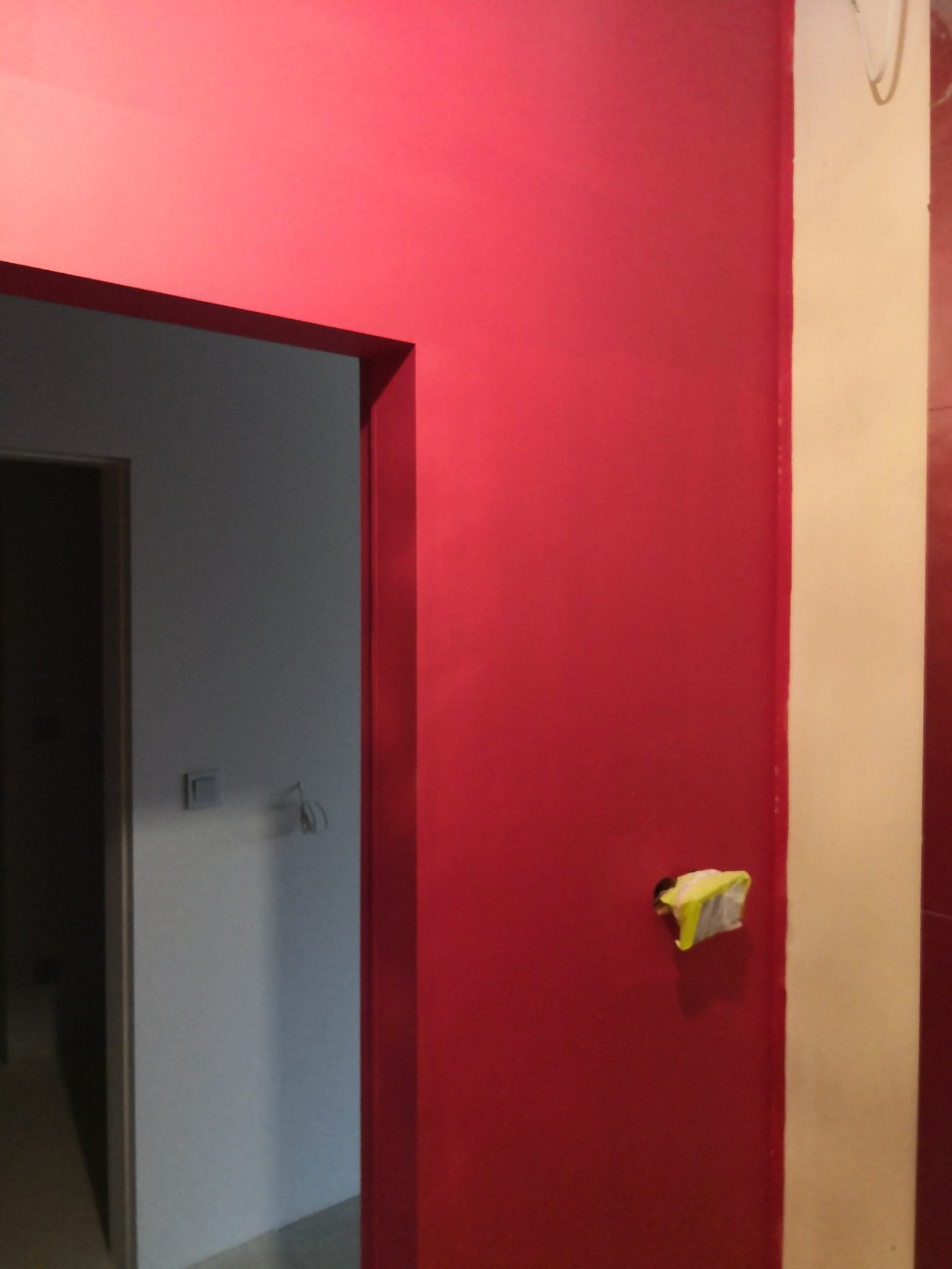Czerwona ściana po malowaniu, widoczny fragment futryny drzwi i zabezpieczone gniazdko. Świeżo pomalowane wnętrze z intensywnym kolorem.