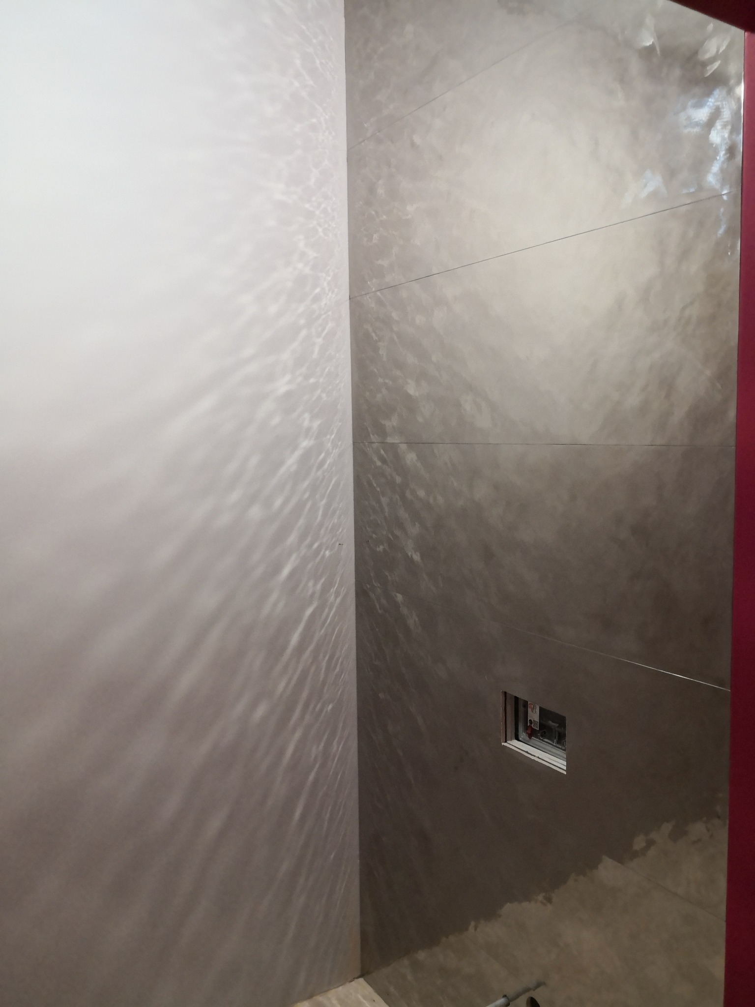 Ściana w łazience z dekoracyjną, metaliczną gładzią w odcieniach szarości, imitującą efekt wody. Widoczna instalacja podtynkowa i fragment czerwonej ramy.