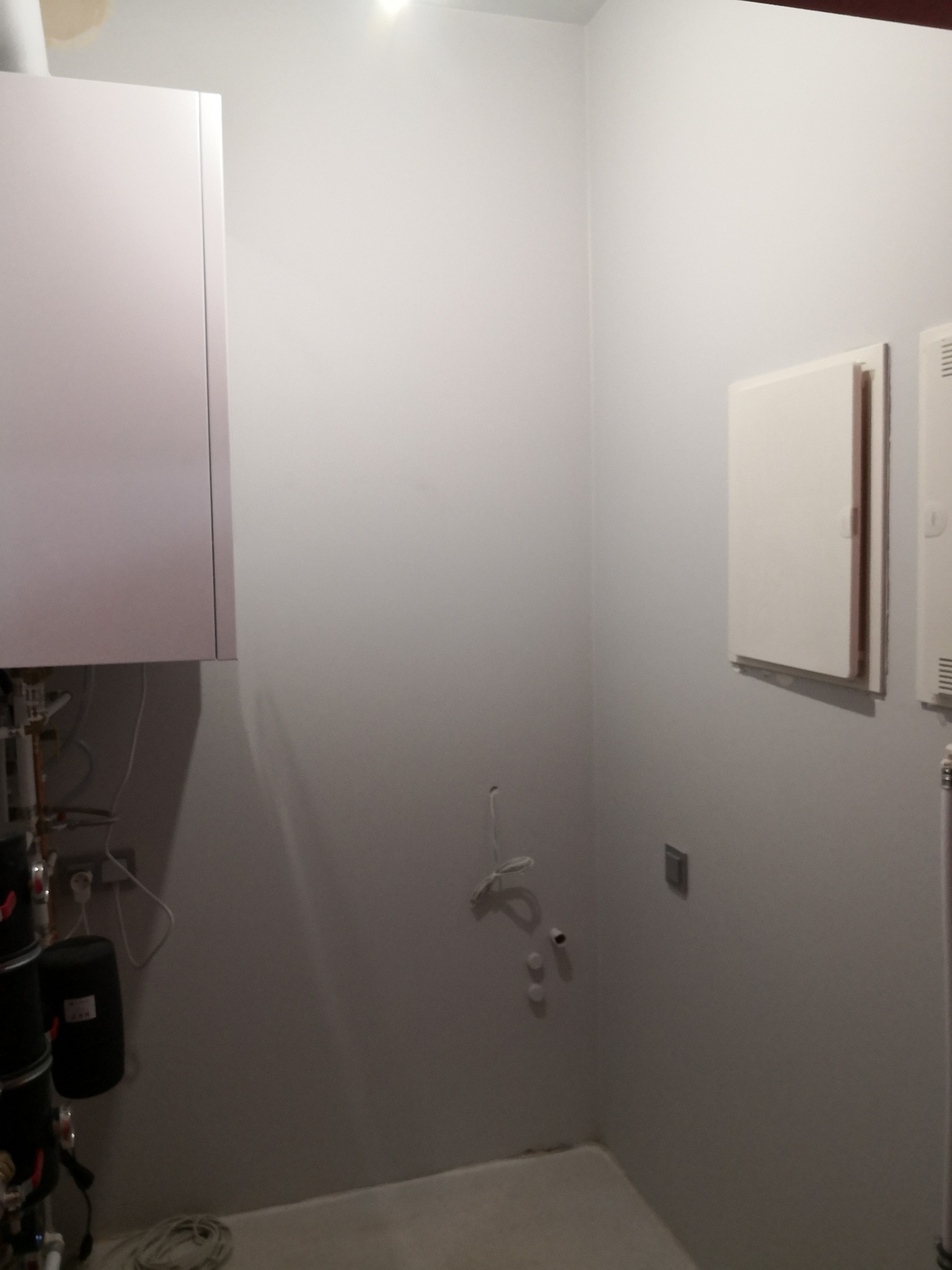 Szare wnętrze kotłowni z białym piecem, instalacją hydrauliczną i grzejnikiem. Widoczne przyłącza na ścianie i kabel na podłodze. Minimalistyczny design.