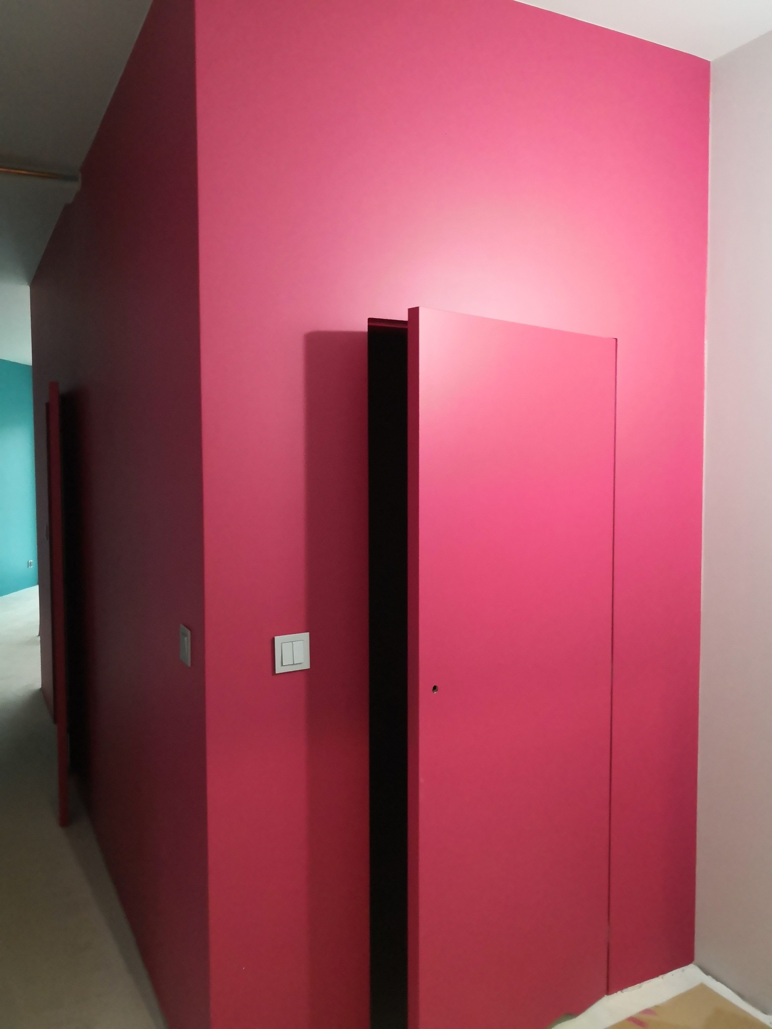 Różowa ściana z ukrytymi drzwiami i włącznikiem światła. Minimalistyczny design wnętrza, świeżo po malowaniu. Widoczny fragment podłogi zabezpieczony folią.