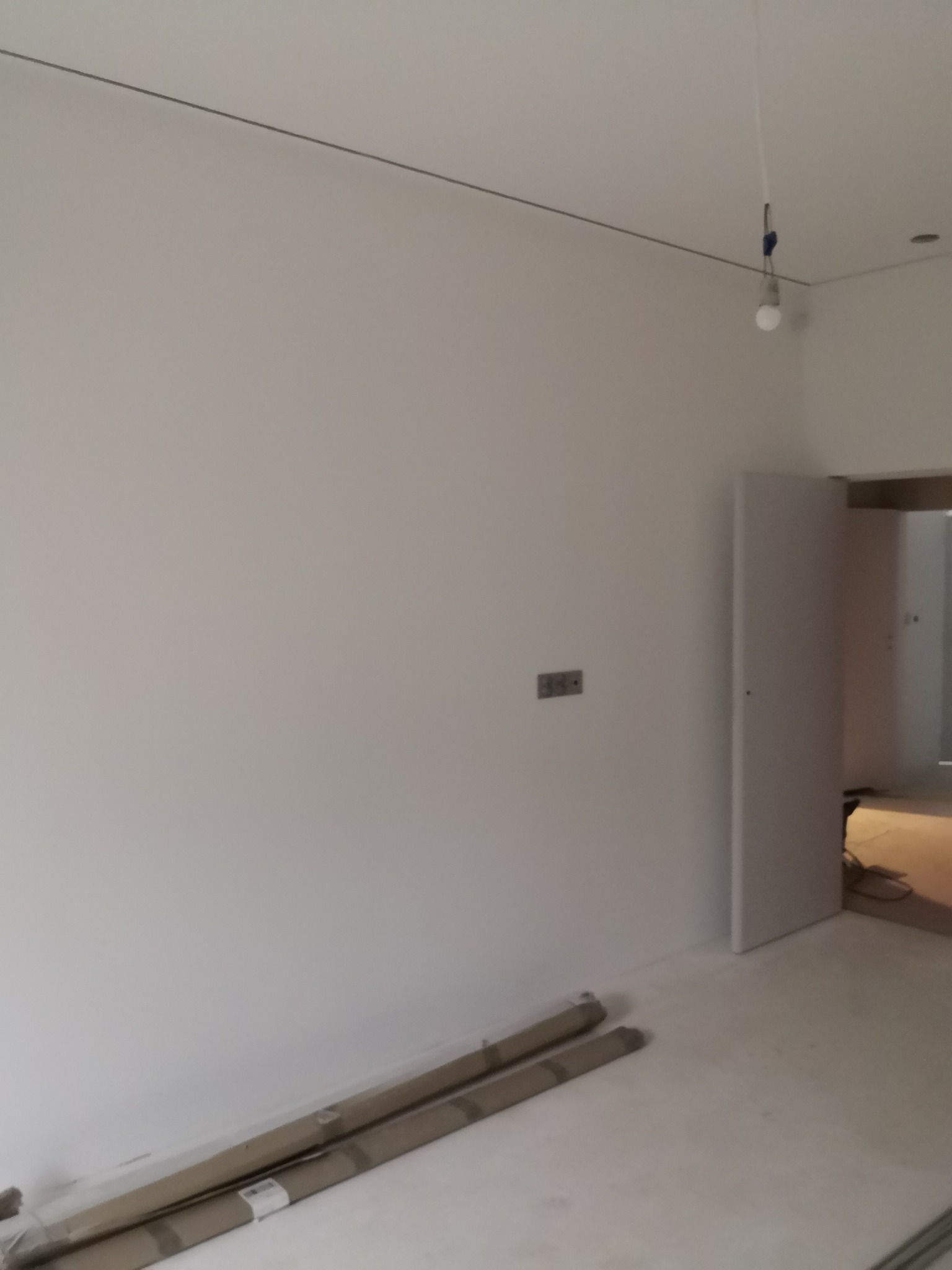 Świeżo pomalowane, białe wnętrze z widocznymi rolkami malarskimi przy ścianie i otwartymi drzwiami do kolejnego pomieszczenia. Minimalistyczny wystrój, surowy stan wykończenia.