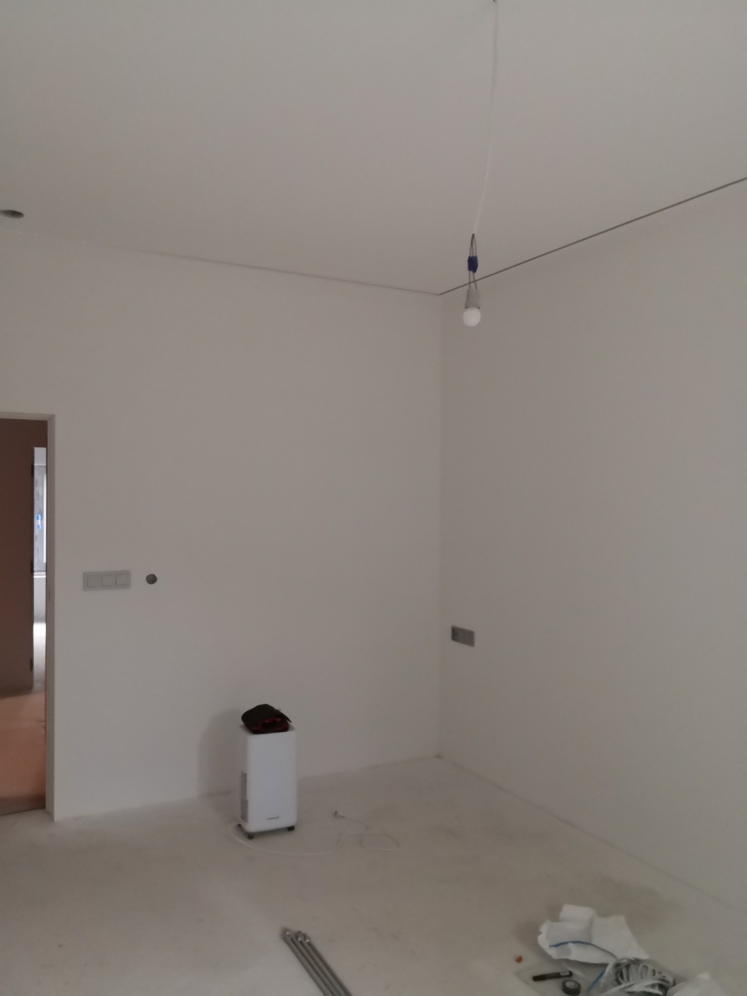 Wnętrze pokoju po malowaniu ścian na biało, widoczny oczyszczacz powietrza, surowa żarówka i narzędzia malarskie na podłodze. Świeżo pomalowane ściany.