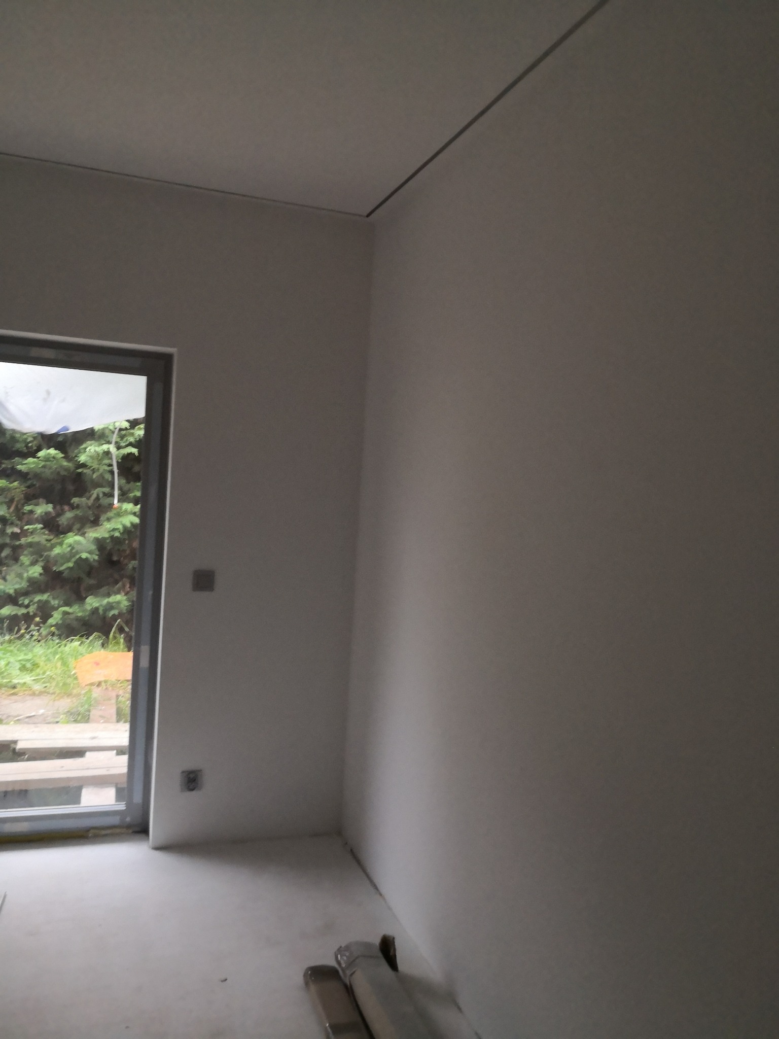 Wnętrze po gładziowaniu ścian i sufitu, widoczne puszki elektryczne, okno z widokiem na zieleń. Na podłodze folia malarska.