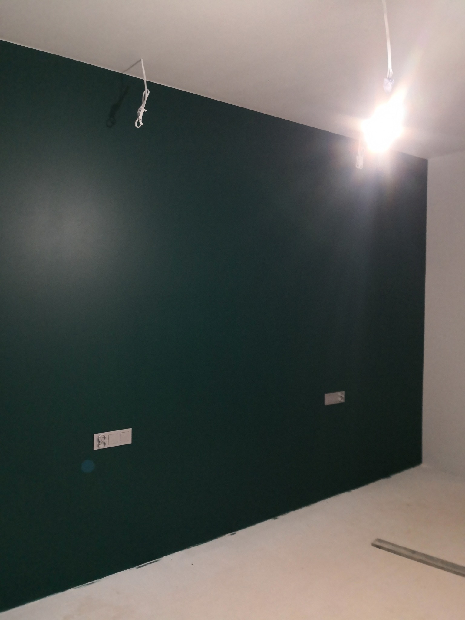Ściana pomalowana na ciemnozielony kolor, widoczne gniazdka elektryczne i przewody pod sufitem. Świeżo pomalowane wnętrze z jasną podłogą.