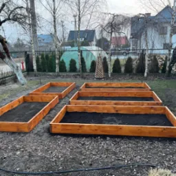 Przygotowane, drewniane skrzynie na podwyższone grządki wypełnione ziemią w ogrodzie, widoczne drzewa i zabudowania w tle.