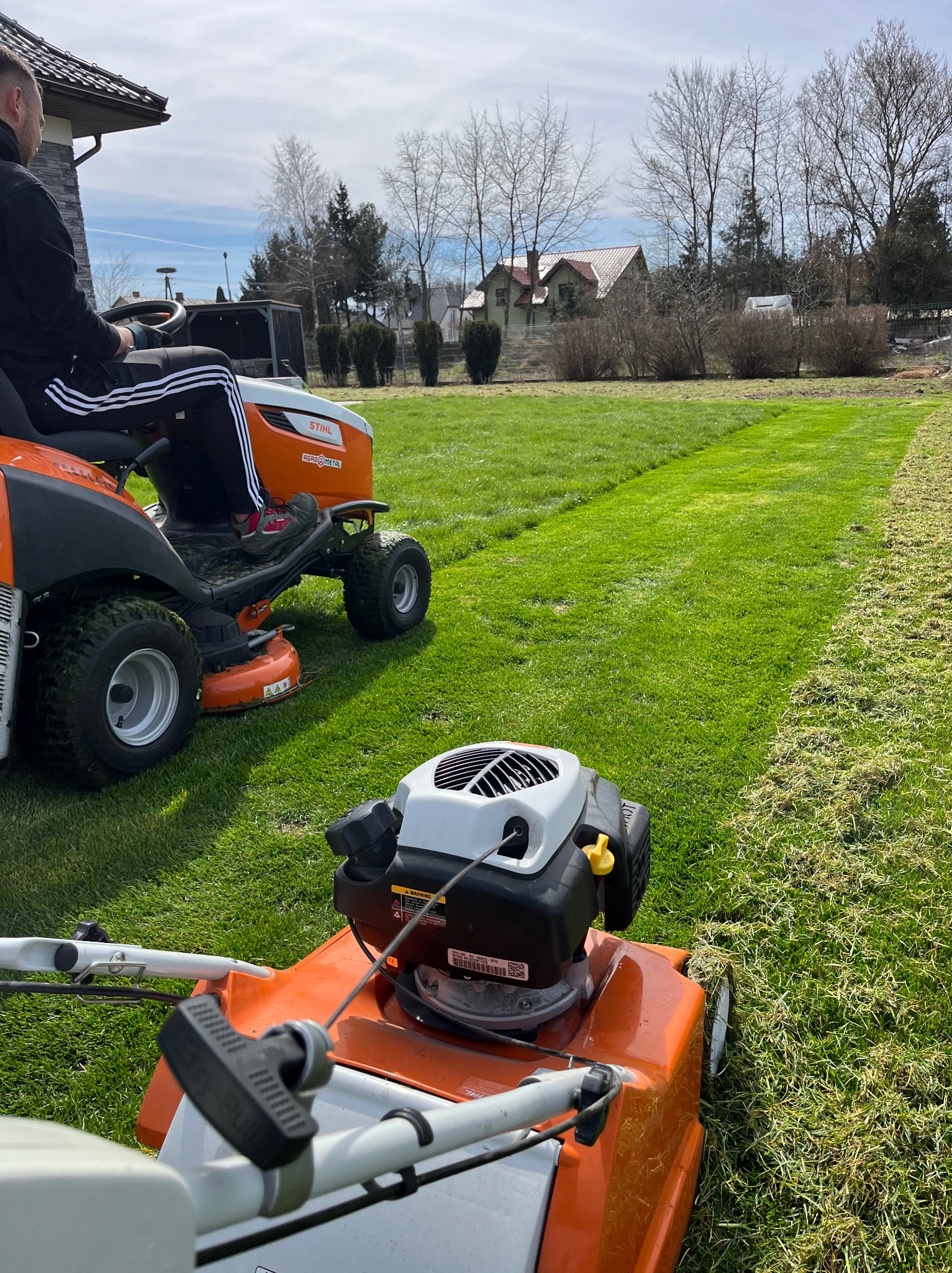 Traktor-kosiarka Stihl i kosiarka spalinowa na trawniku po koszeniu. Widoczny wzór w pasy i fragment domu w tle.