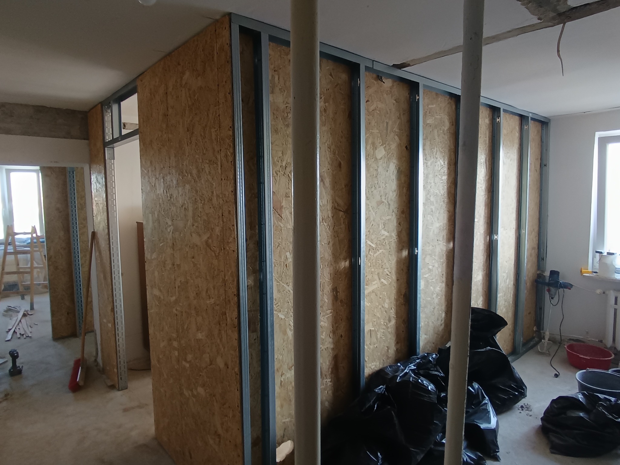 Wnętrze pomieszczenia w trakcie budowy ścianek działowych z profili metalowych i płyt drewnopochodnych, z widocznymi otworami drzwiowymi i rurą instalacyjną.