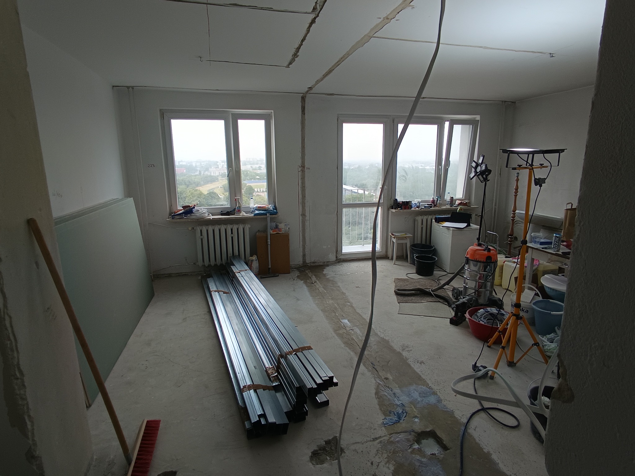Pomieszczenie w trakcie prac wykończeniowych, z ułożonymi na betonowej posadzce metalowymi profilami i płytami budowlanymi. Dwa okna z widokiem na miasto i sprzęt budowlany wskazują na początkowy...
