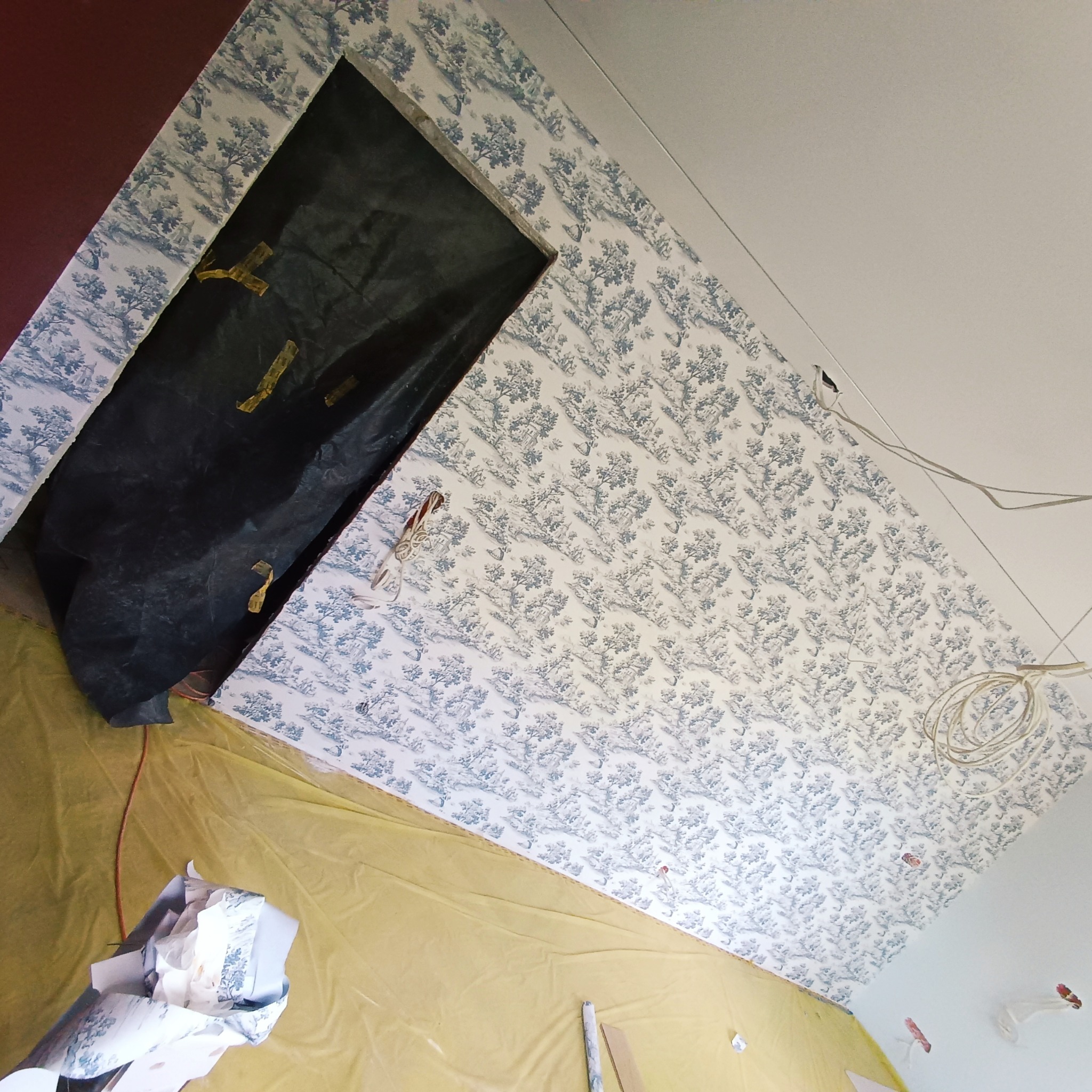 Pokój w trakcie prac wykończeniowych z częściowo położoną tapetą w niebiesko-biały wzór krajobrazowy, zabezpieczoną żółtą folią podłogą i zakrytymi drzwiami.