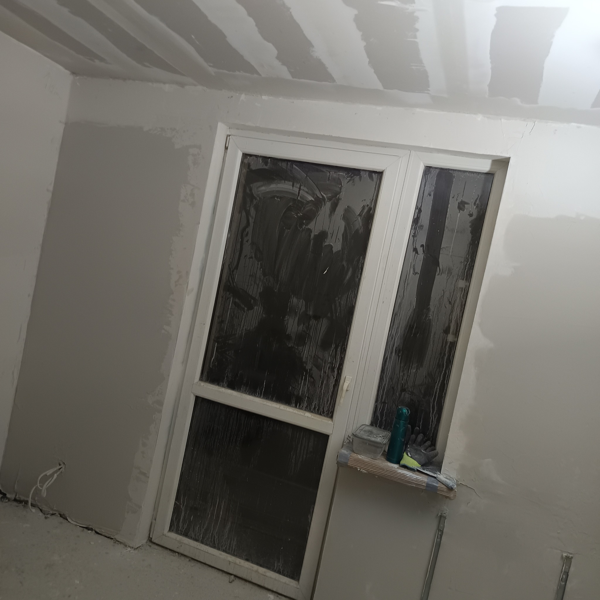 Pomieszczenie w trakcie prac wykończeniowych, z częściowo wyrównanymi ścianami i sufitem, gdzie widoczne są pasy łączeń. Białe okno balkonowe z zabrudzoną szybą, na parapecie narzędzia budowlane...