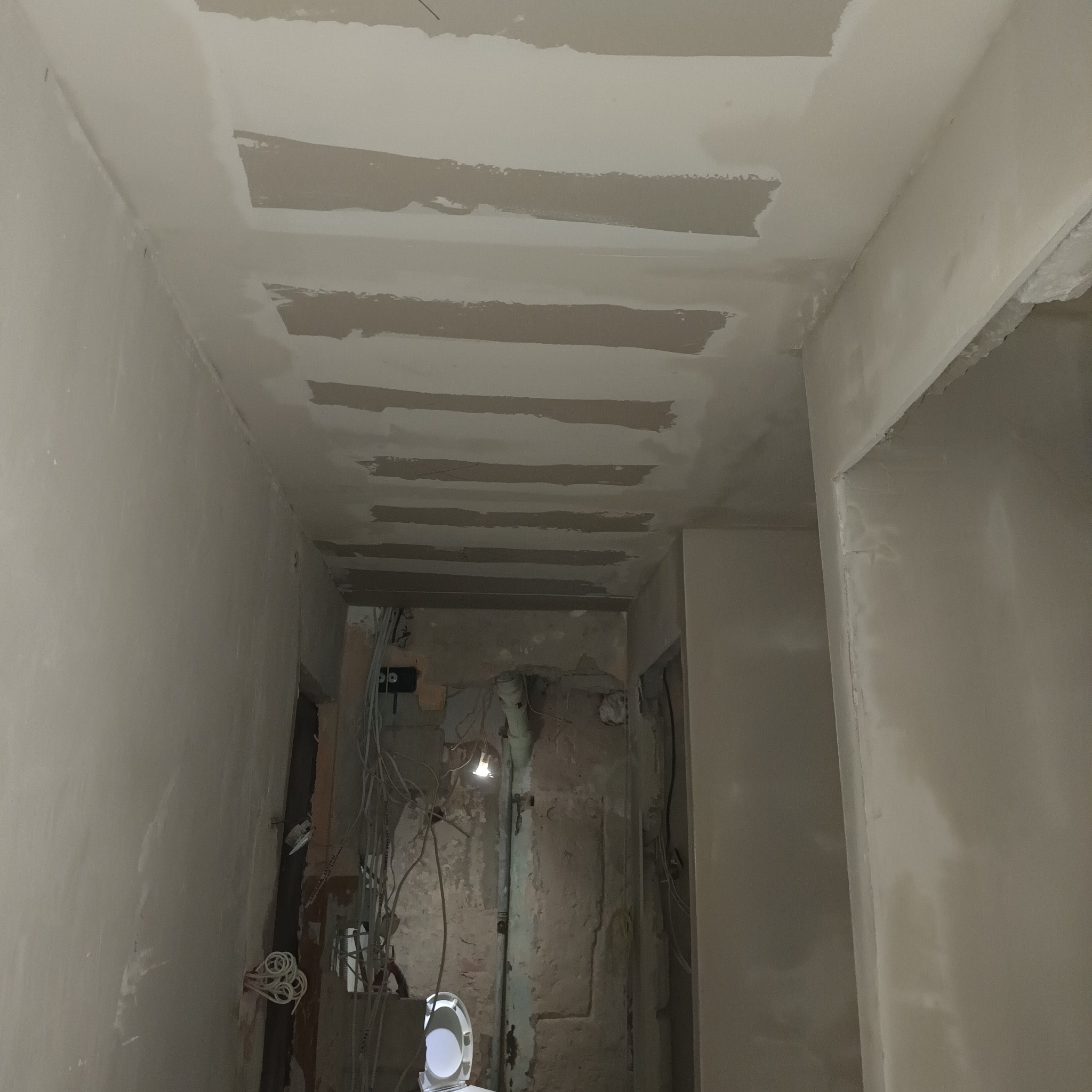 Sufit w korytarzu z wyraźnymi pasami świeżej gładzi, ściany częściowo wykończone, a w głębi pomieszczenia widoczne surowe instalacje i rury.
