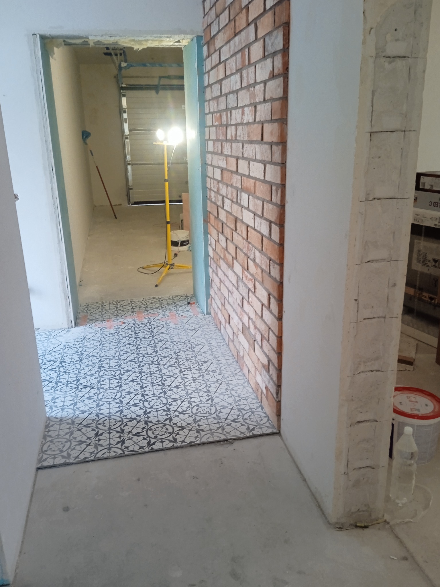 Wnętrze w trakcie remontu: dekoracyjne płytki podłogowe w geometryczny wzór, fragment ściany wyłożonej cegłą klinkierową, lampa budowlana w tle.