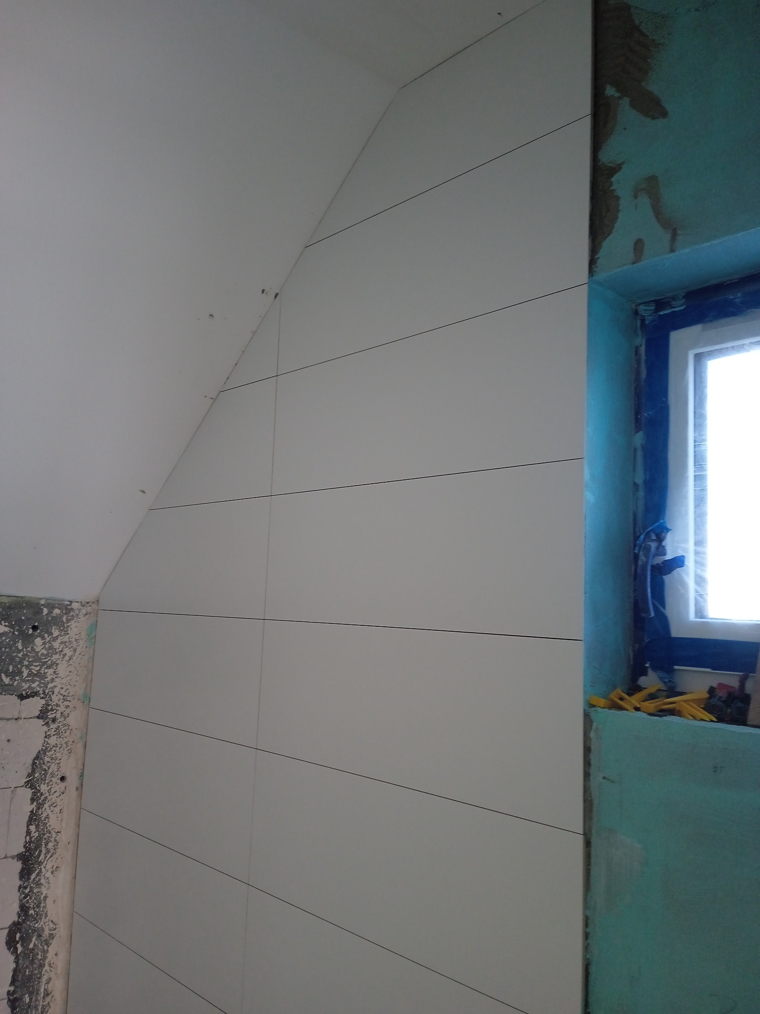 Ściana w łazience wyłożona białymi płytkami z ciemnymi fugami, widoczne przygotowania wokół okna i surowy fragment ściany w rogu.