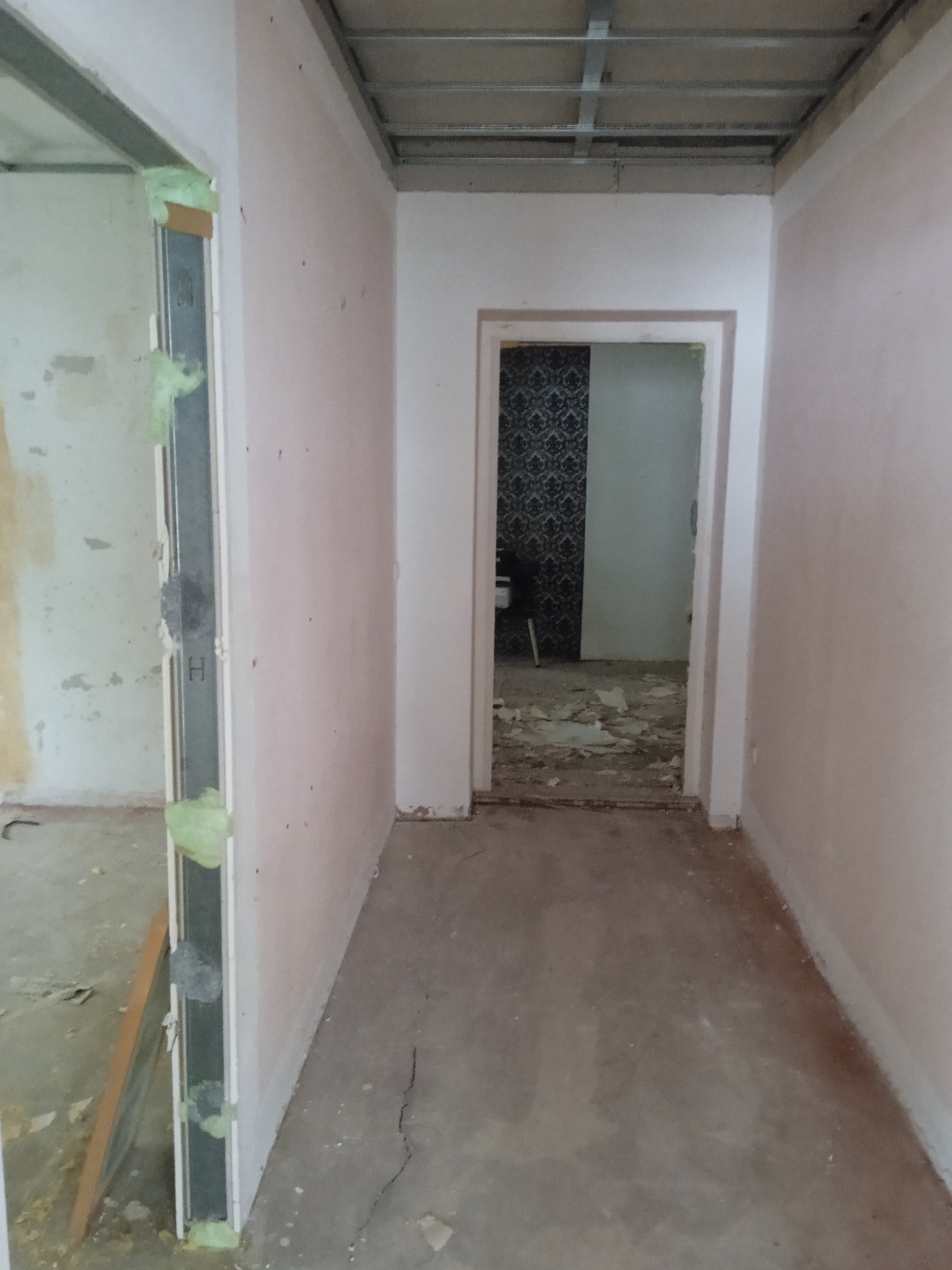 Korytarz w trakcie prac: surowy beton na podłodze, ściany w jasnym odcieniu różu, widoczne profile sufitowe i fragmenty pianki montażowej przy futrynach.