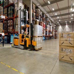 KMC-Services - Wnętrze magazynu z wysokimi regałami wypełnionymi towarem, wózek widłowy transportuje paletę z zapakowanymi produktami, na podłodze widoczne żółte linie wyznaczające strefy.