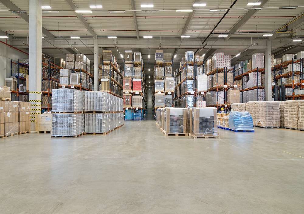 Pełny magazyn z rzędami zapakowanych i zabezpieczonych folią towarów na paletach, umieszczonych na wysokich metalowych regałach.