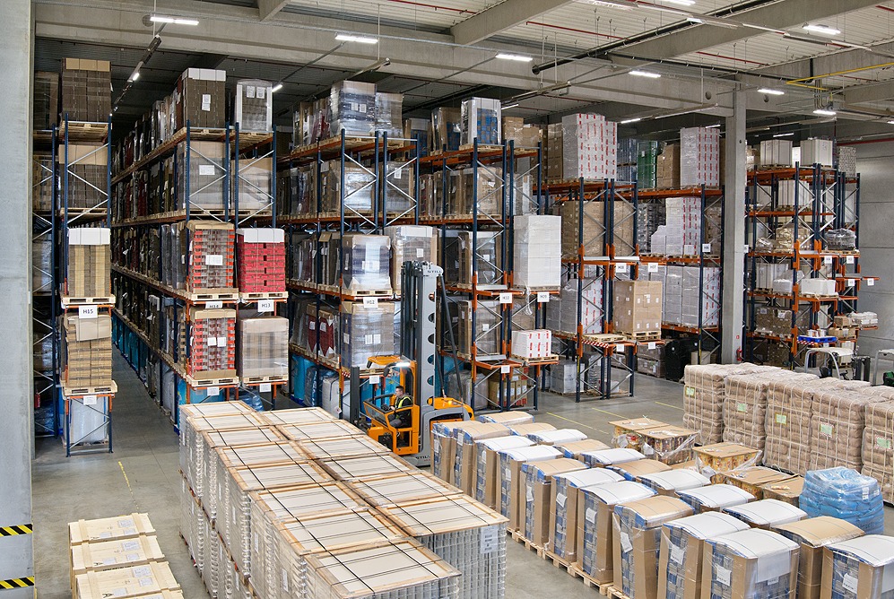 Wnętrze magazynu wysokiego składowania z regałami wypełnionymi paletami z towarem, wózek widłowy w trakcie pracy.