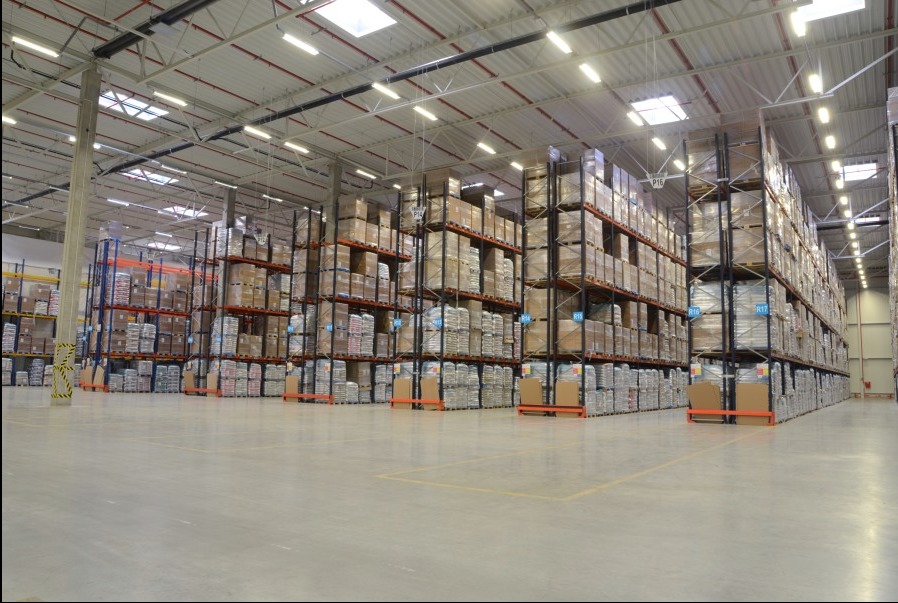 Wnętrze magazynu wysokiego składowania z regałami wypełnionymi kartonami i workami, widoczne oznaczenia regałów P16, R13, R16, R17.