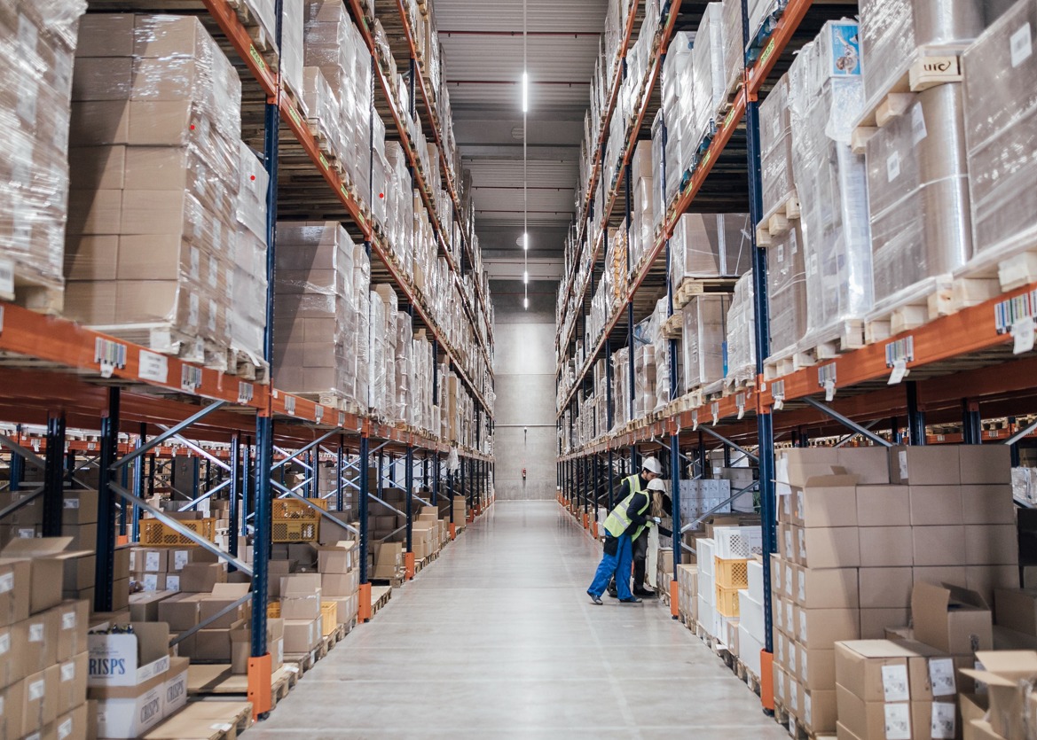Długi korytarz magazynu z wysokimi regałami wypełnionymi kartonami i folią, dwóch pracowników w kaskach i kamizelkach odblaskowych pochylonych nad paczkami na podłodze.