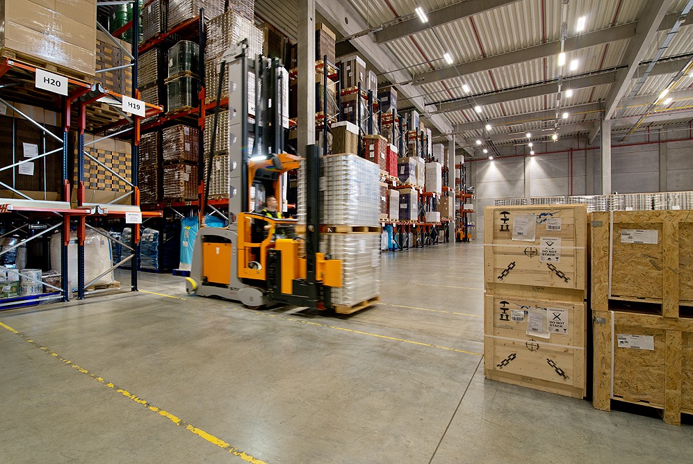 Wnętrze magazynu z wysokimi regałami wypełnionymi towarem, wózek widłowy transportuje paletę z zapakowanymi produktami, na podłodze widoczne żółte linie wyznaczające strefy.