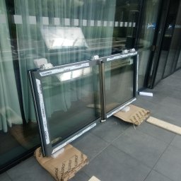 NI-TECH ROMAN Jedenasty - Dwa okna aluminiowe marki Aluprof, oparte o kartony i drewniane deski, czekają na montaż w budynku z widocznymi szklanymi fasadami i logo Radisson.