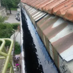 Rynna dachowa wypełniona wodą, widok z góry z drabiny, dach pokryty czerwoną dachówką, w tle budynki mieszkalne i zieleń.