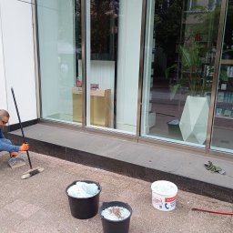 NI-TECH ROMAN Jedenasty - Mężczyzna w pomarańczowych rękawicach sprząta potłuczone szkło przed witryną sklepową, używając zmiotki i szufelki; obok stoją wiadra wypełnione szkłem i puste opakowanie po zaprawie.